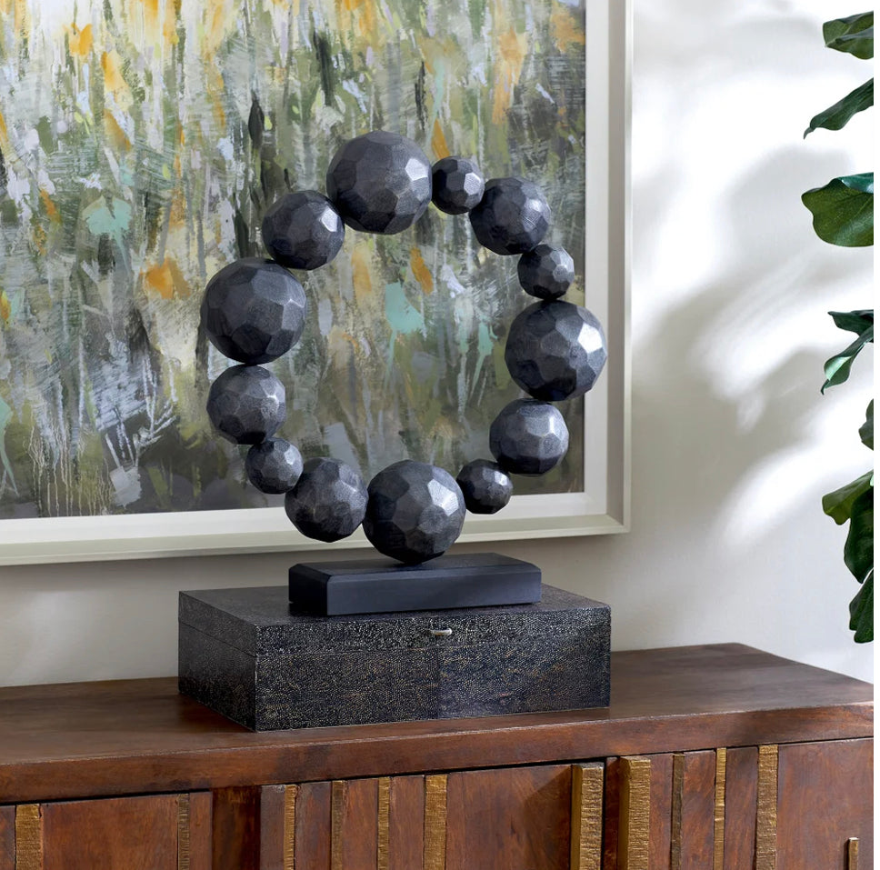 The chiseled spheres of the Garnet sculpture connect to form a circle perched atop a solid wood base. Made from Ardu wood and bearing a black washed finish, the sculpture serves as an enchanting addition to any setting. By displaying the Garnet sculpture, you are sure to fascinate your guests with its simplistic charm.  Artisan Produced: This product, or portions of this product have been hand crafted by artisans. Each piece is unique and can vary in finish, tone, texture and color.  Dimensions: L 17.5 X W 